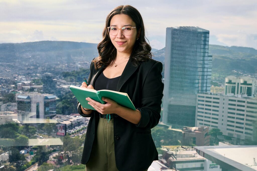 Woman holding a notebook in a modern office environment, standing near a desk and looking confidently toward the camera.
